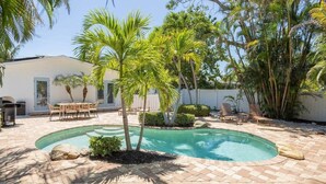 Zona de piscina con palmeras, un patio con sillas y un edificio blanco al fondo.