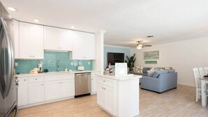 Una cocina moderna con armarios blancos, lavavajillas y un revoque de azulejos azules. Cuenta con una zona de comedor con un sofá gris y un ventilador de techo.