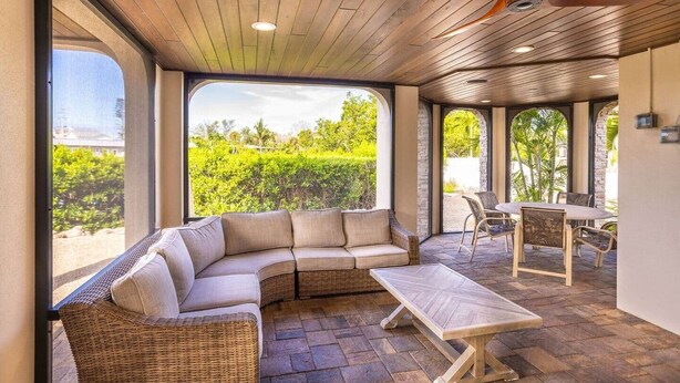 Ein überdachter Patio-Bereich mit einer gebogenen Couch, einem Esstisch und Stühlen, mit großen Fenstern und Blick auf Grünflächen.