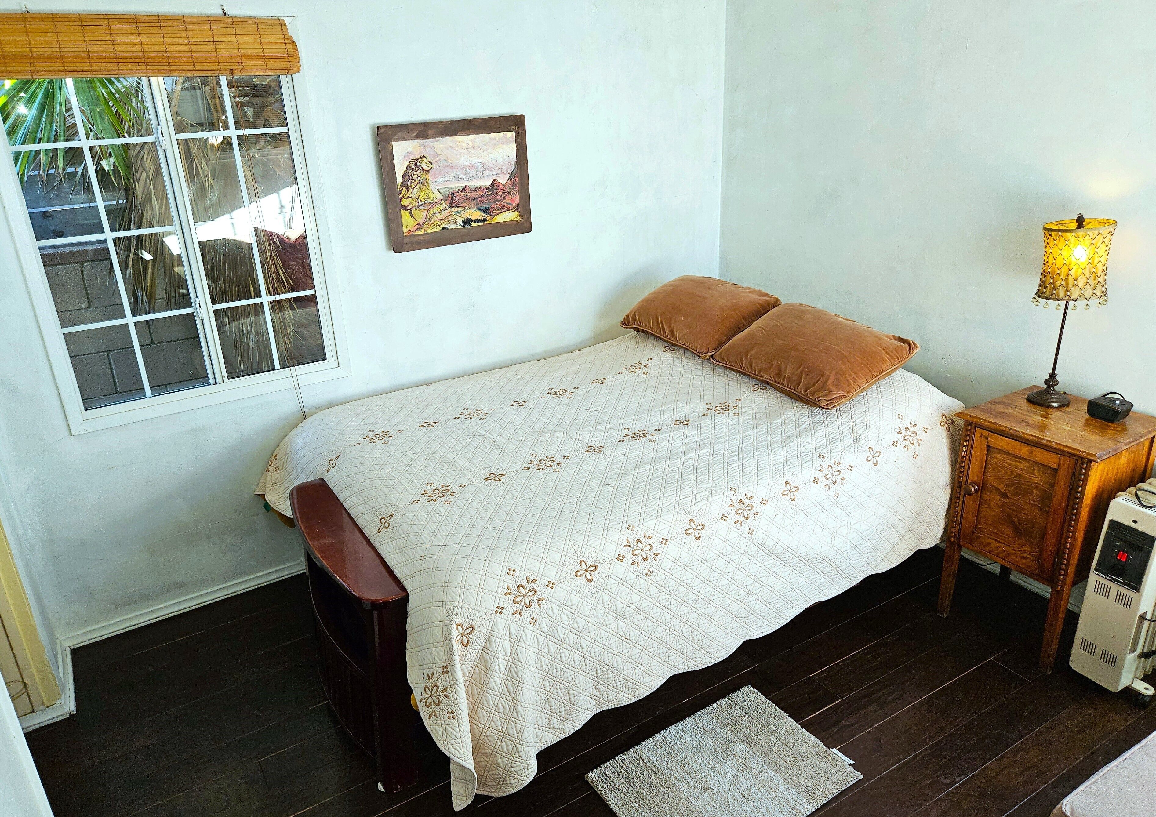 A bedroom with a bed, a nightstand, a lamp, a heater, and a window with blinds.