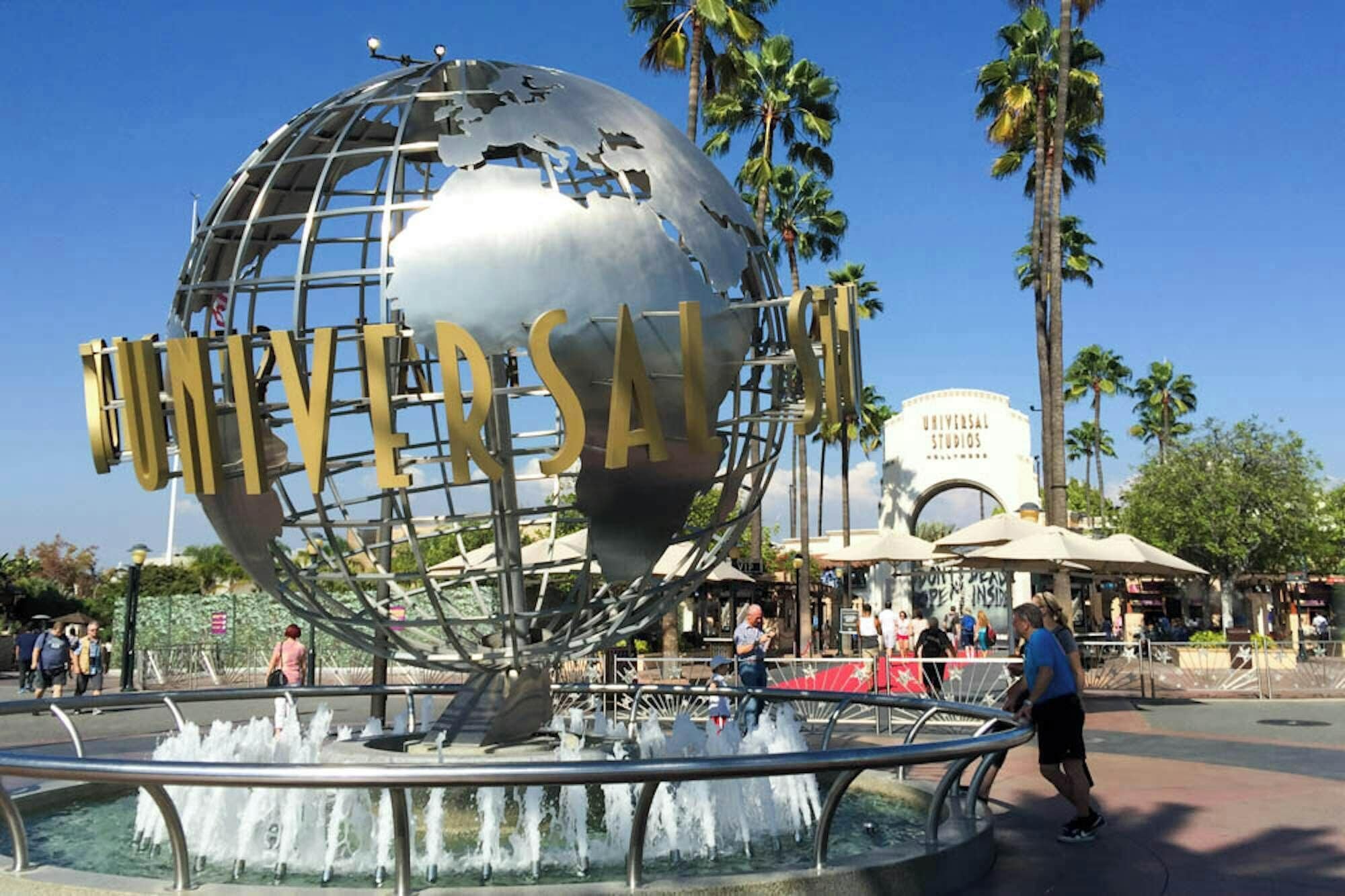 Un globo con la palabra "UNIVERSAL" escrita, rodeado por una fuente de agua, con palmeras y el edificio de Universal Studios al fondo.
