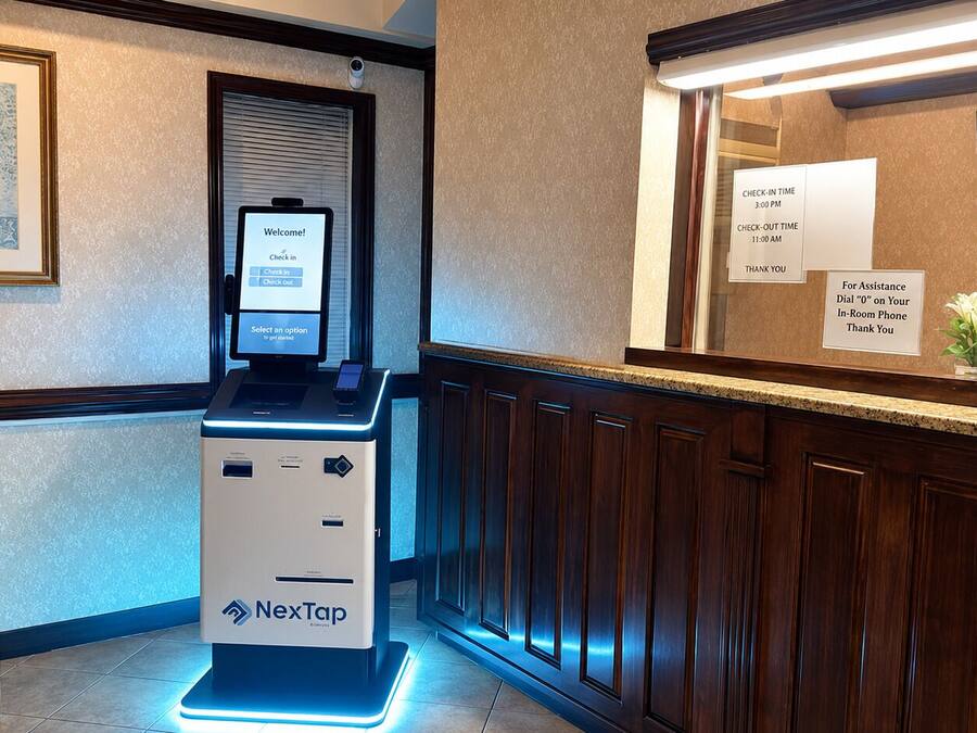 A NexTap self-service kiosk in a hotel lobby with a check-in/check-out board, a framed picture on the wall, and a small table with a plant.