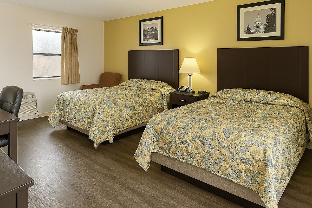 Ein Hotelzimmer mit zwei Betten, einem Schreibtisch, einem Stuhl, einer Lampe und zwei gerahmten Bildern an der Wand.