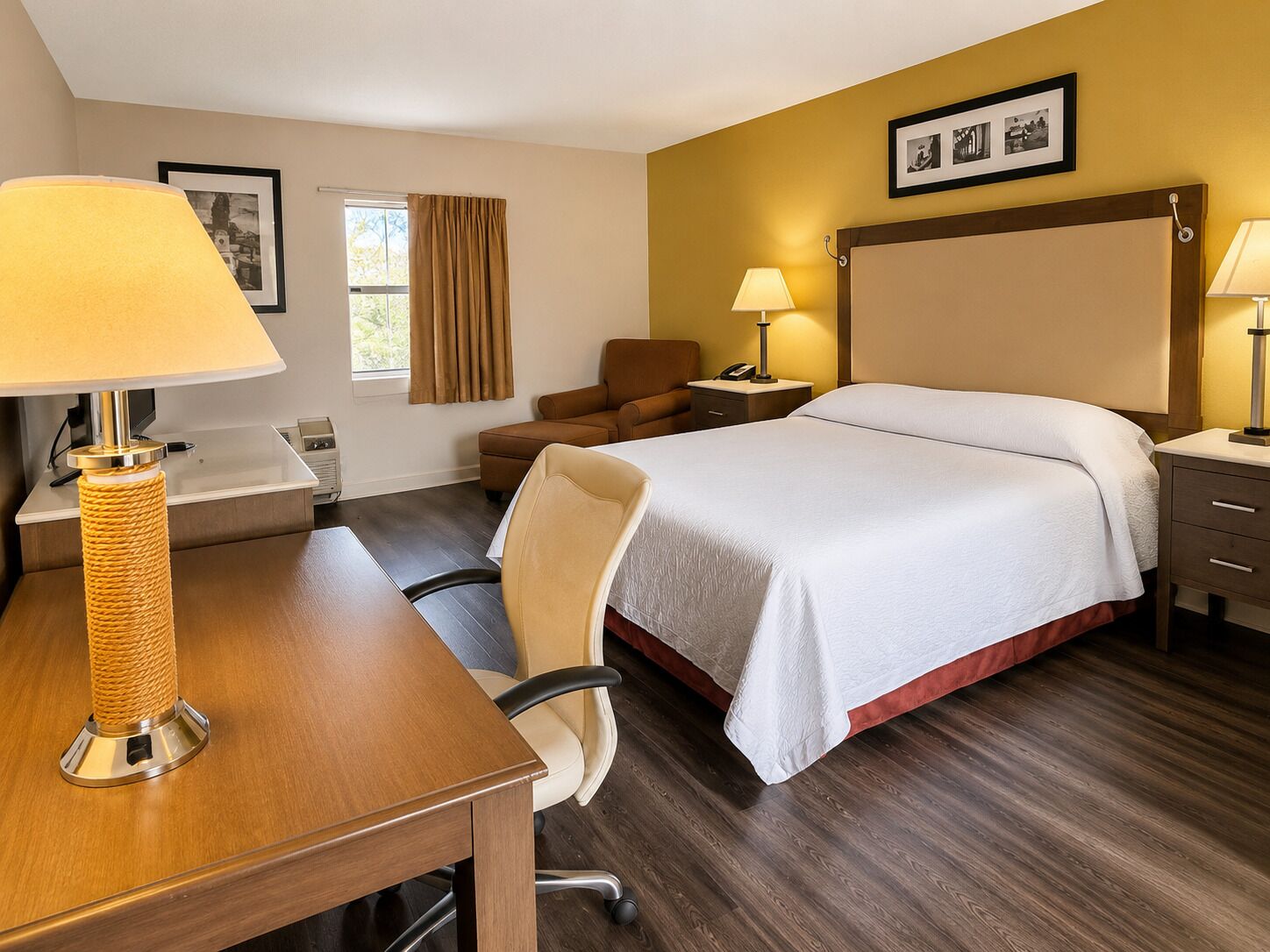 Une chambre d’hôtel comprenant un lit, un bureau avec une lampe, une chaise, une table de chevet, une commode et une fenêtre avec des rideaux.