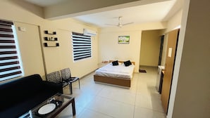 A hotel room with a bed, a desk with a chair, a window with blinds, a ceiling fan, and a door leading to another room. - Regal Suites (Thiruvananthapuram)