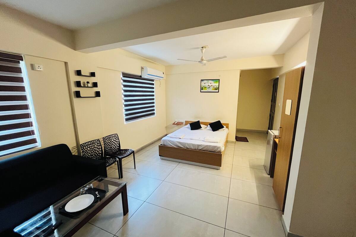 A hotel room with a bed, a desk with a chair, a window with blinds, a ceiling fan, and a door leading to another room.