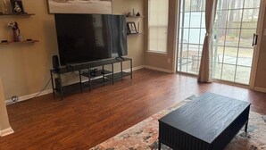 A living room with hardwood flooring, a black entertainment unit, a large flat-screen TV, and a dark coffee table. - Come Stay At The Orchard's Inn, 15 minutes away from Airport! (Summerville)