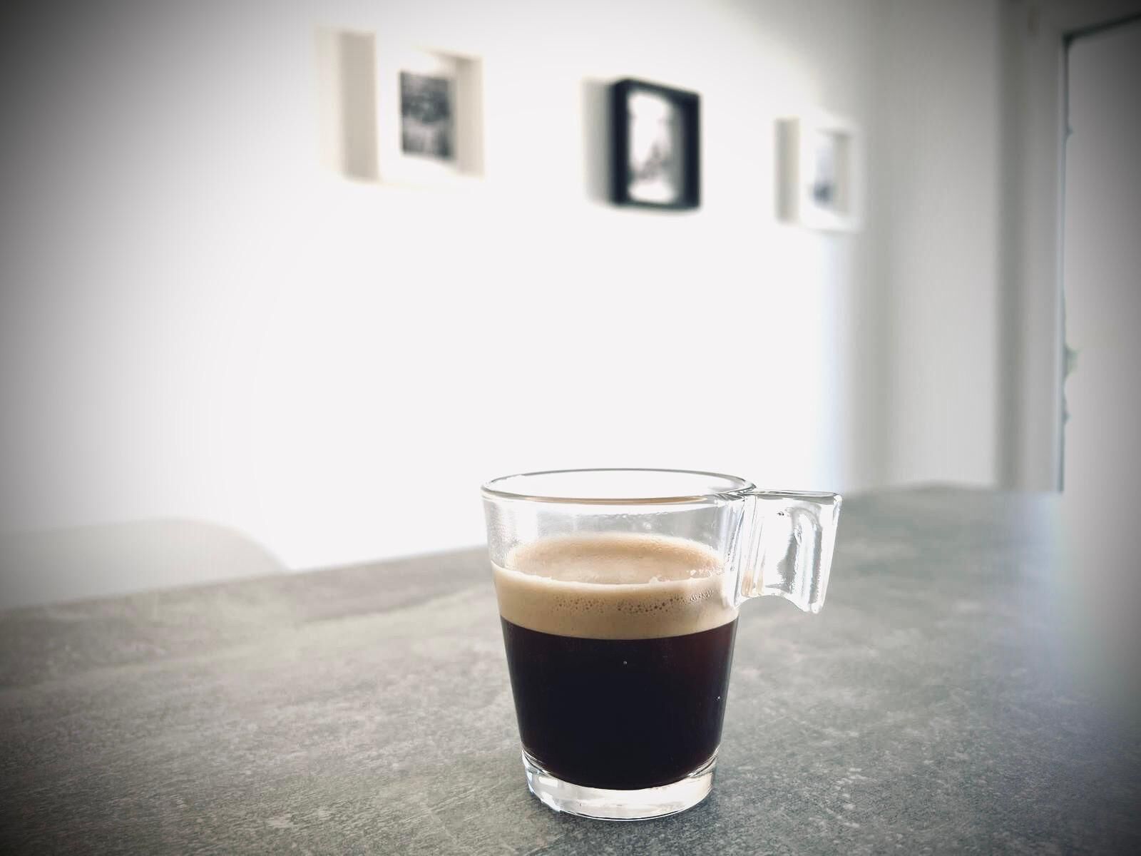 Une tasse en verre remplie d’un liquide sombre, probablement du café, posée sur une surface texturée, avec un arrière-plan flou où l’on aperçoit des photos encadrées accrochées au mur.