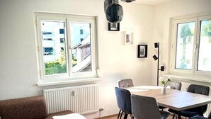 Uma sala de estar moderna com um sofá, uma mesa de centro de vidro, uma mesa de jantar com cadeiras e vista para o exterior através de amplas janelas.