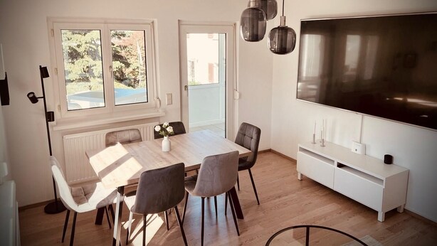 Uma sala de jantar com uma mesa grande, quatro cadeiras, um vaso com flores, luminárias pendentes, uma TV de tela plana grande e um armário branco.