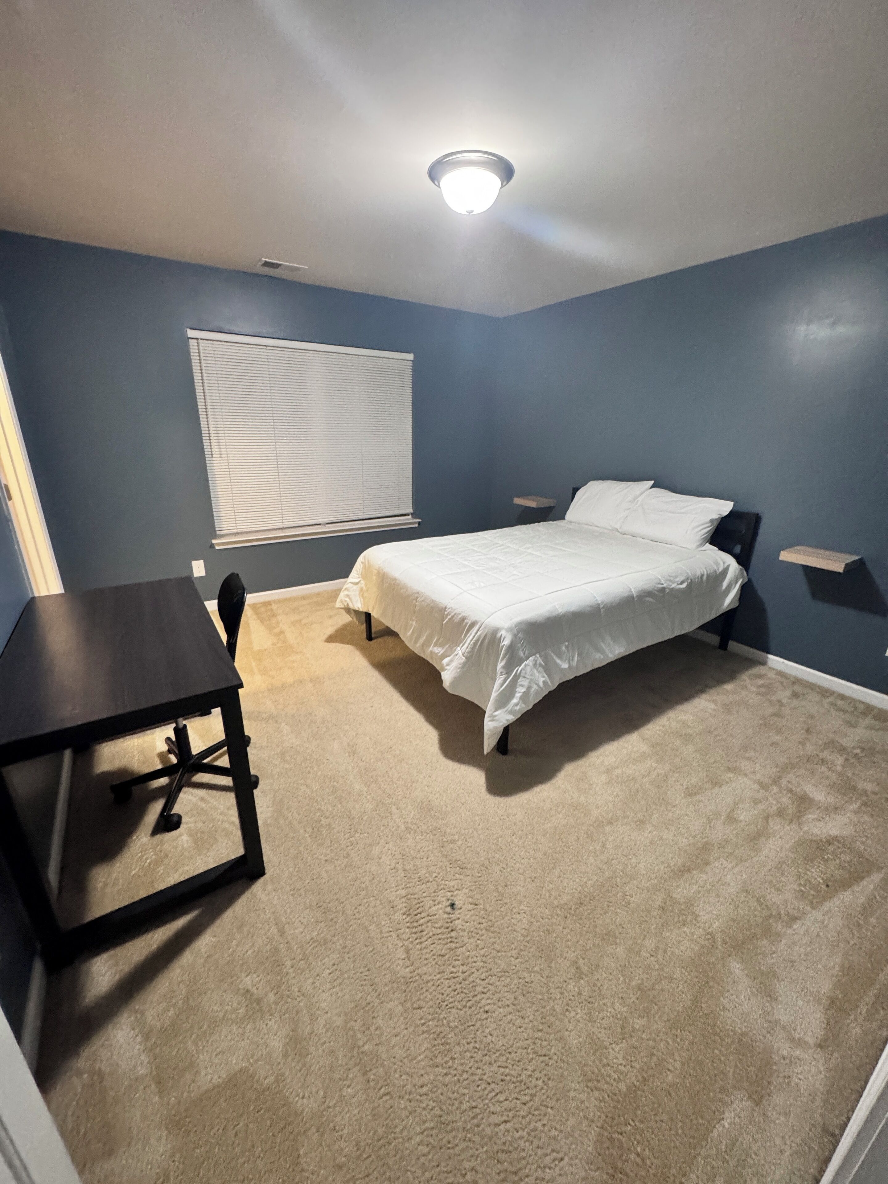 A bedroom with a bed, a desk, and a window with blinds.