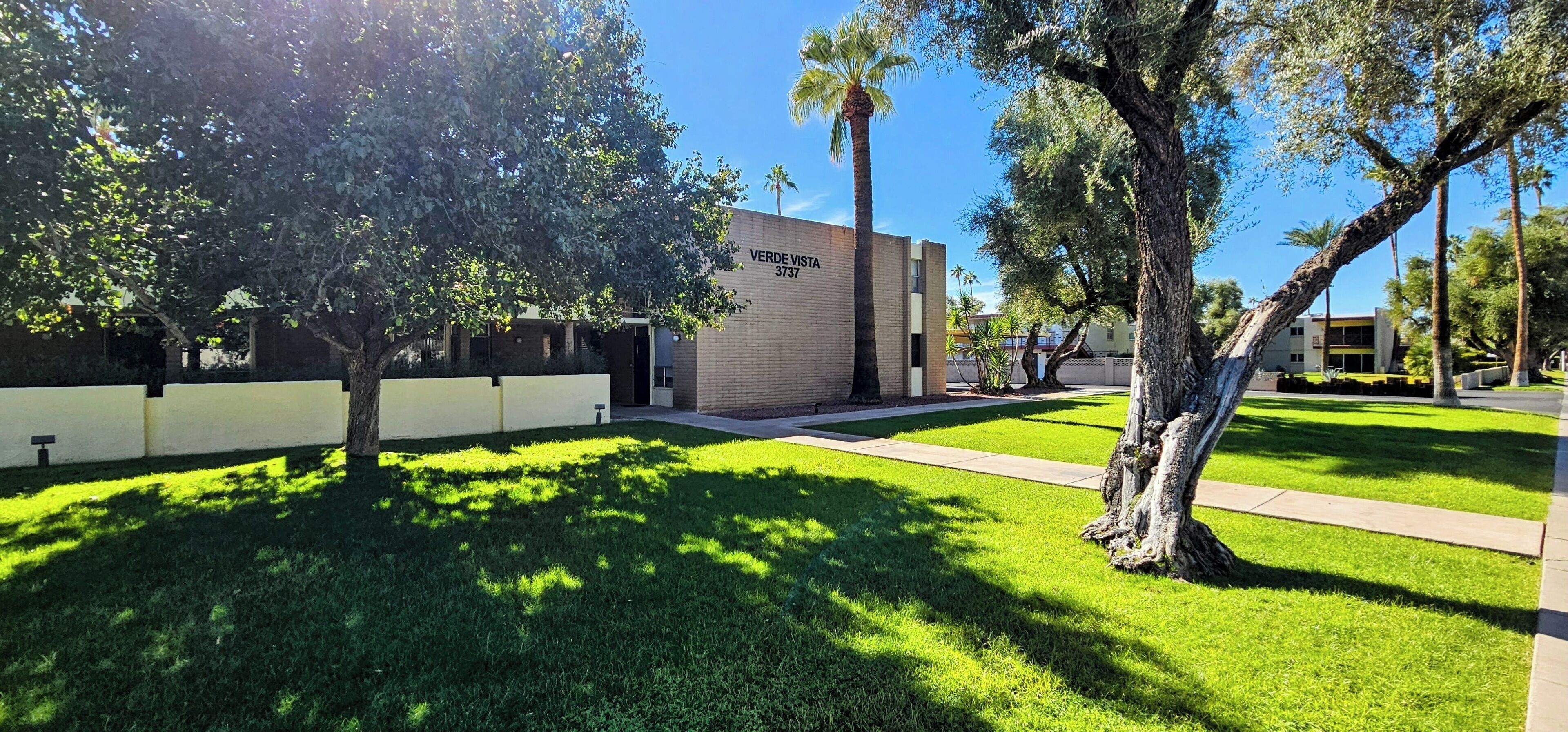 A building with a sign reading "VERDE VISTA 3737" in a well-maintained grassy area with trees and a clear sky.