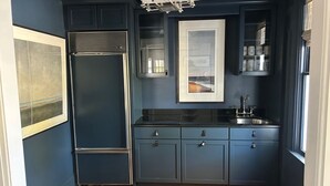 Une cuisine compacte avec des meubles de couleur bleu foncé, un réfrigérateur en acier inoxydable, un four encastré et une fenêtre équipée de stores.