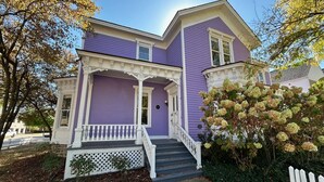 Une maison à deux étages, à l’extérieur violet et aux ornements blancs, dotée d’un porche avec une balustrade blanche et d’un petit escalier menant à l’entrée.