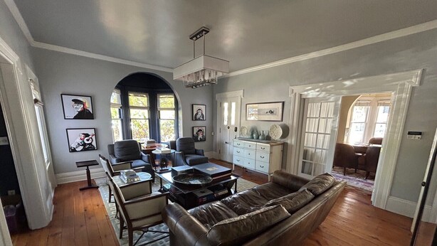 Un salon spacieux avec un canapé en cuir, des fauteuils et une table basse. Les murs sont ornés de tableaux encadrés et un lustre est suspendu au plafond.