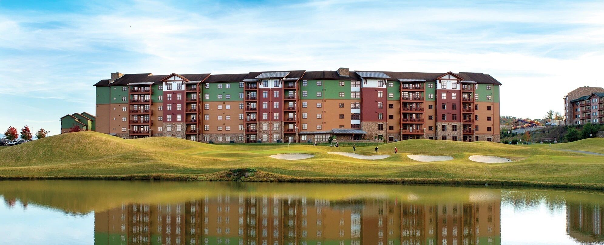 A multi-story hotel with a reflective pool in front, surrounded by green hills and a clear sky.