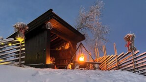 A wooden cabin with a visible fireplace, surrounded by snow-covered trees and a wooden fence.