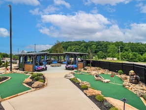 Um campo de golfe com dois gazebos de madeira, um caminho central e um cenário florestal.