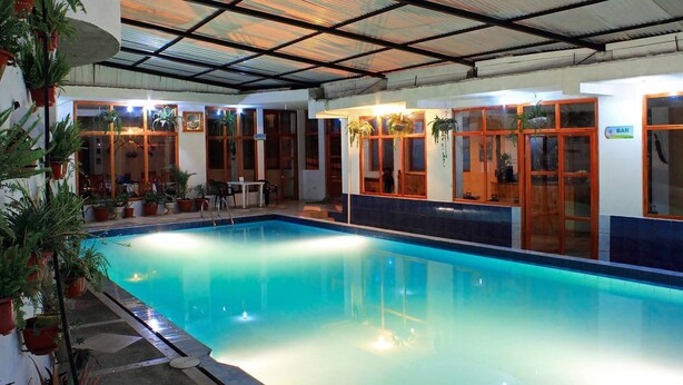 An indoor swimming pool with a tiled floor, surrounded by potted plants and wooden doors. - Hostal y Complejo Turistico La Rueda (Guaranda)