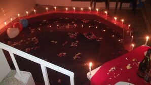 A circular arrangement of candles and rose petals on a dark floor, with balloons and a table with a bottle and glass. - Hostal y Complejo Turistico La Rueda (Guaranda)