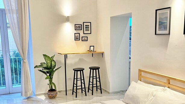 Une chambre à coucher moderne avec un lit, un bureau, deux tabourets, une plante en pot et des tableaux encadrés accrochés au mur.