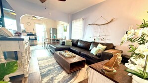 A modern living room with a black leather sofa, a wooden coffee table, and a white cabinet. There is a dining area with a wooden table and chairs, and a kitchen visible in the background. - Stay Local at Your Beachfront Casa in Paradise (Maunabo)
