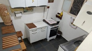 A compact kitchen with white cabinets, a stove, a sink, and a small refrigerator. There are wooden stools and a mat on the floor.