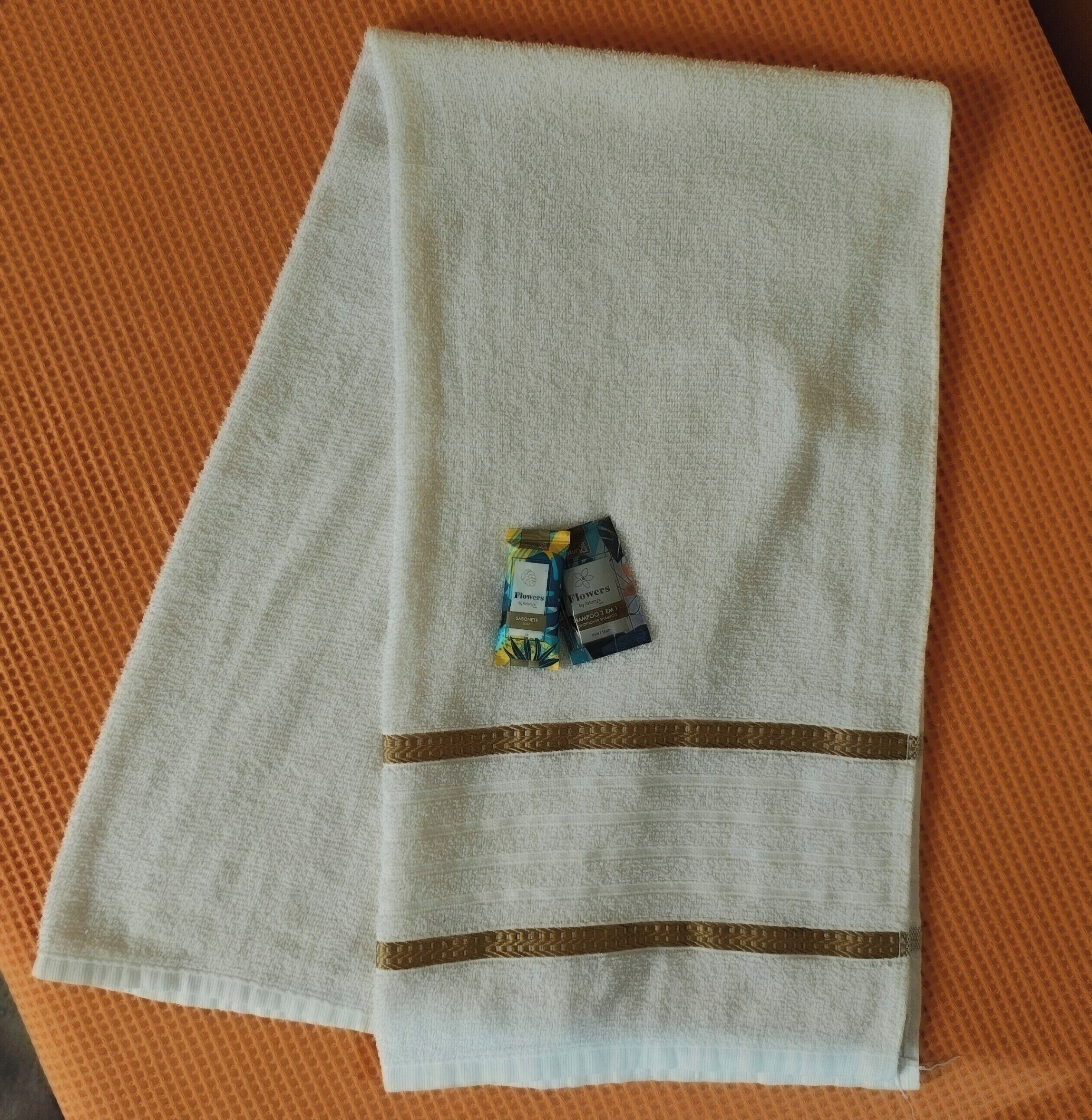 A white towel with a gold stripe and a small packet of shampoo on an orange textured surface.