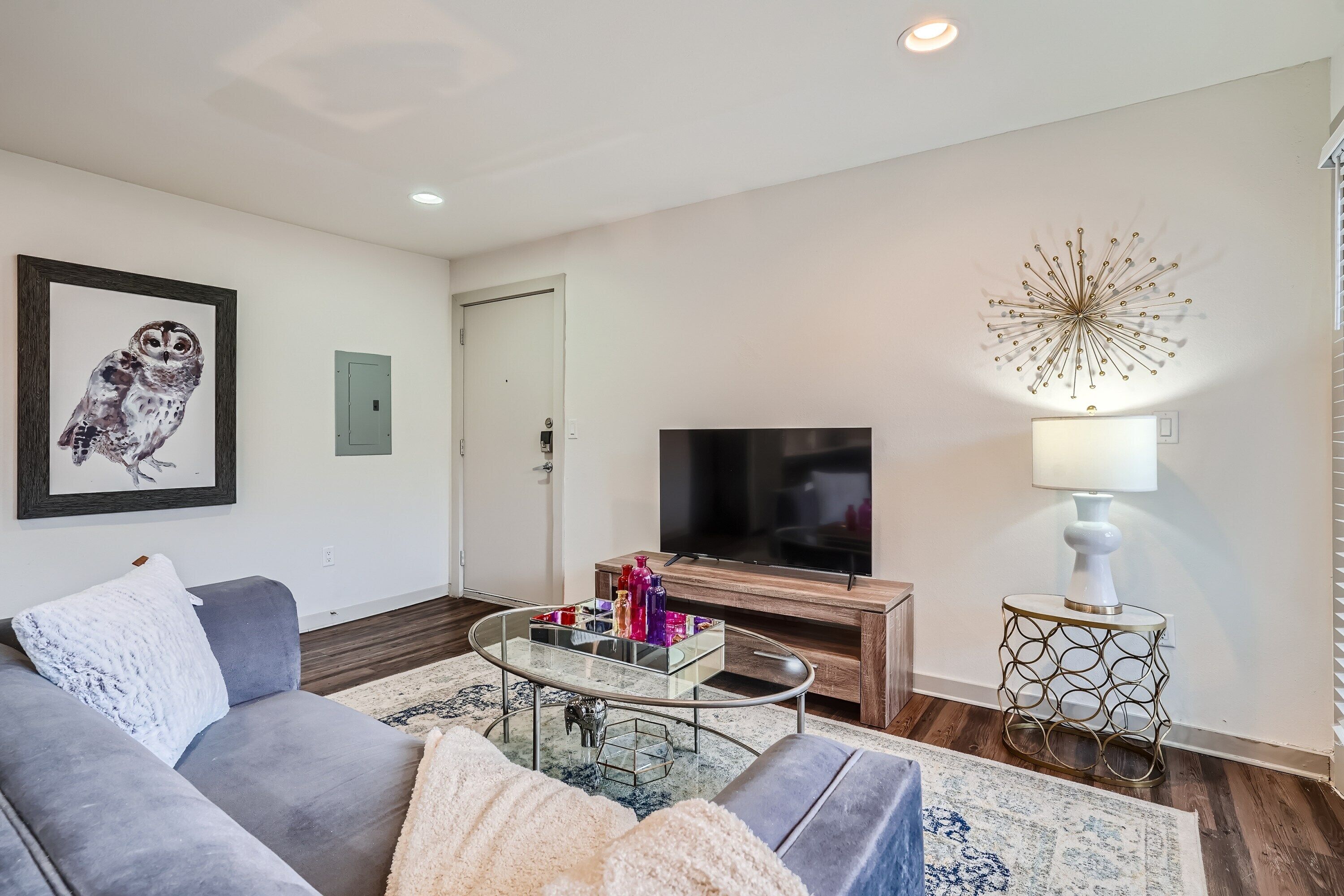 Una sala de estar moderna con un sofá gris, una mesa de centro de vidrio, un televisor de pantalla plana, un elemento decorativo en la pared y un tapete estampado.