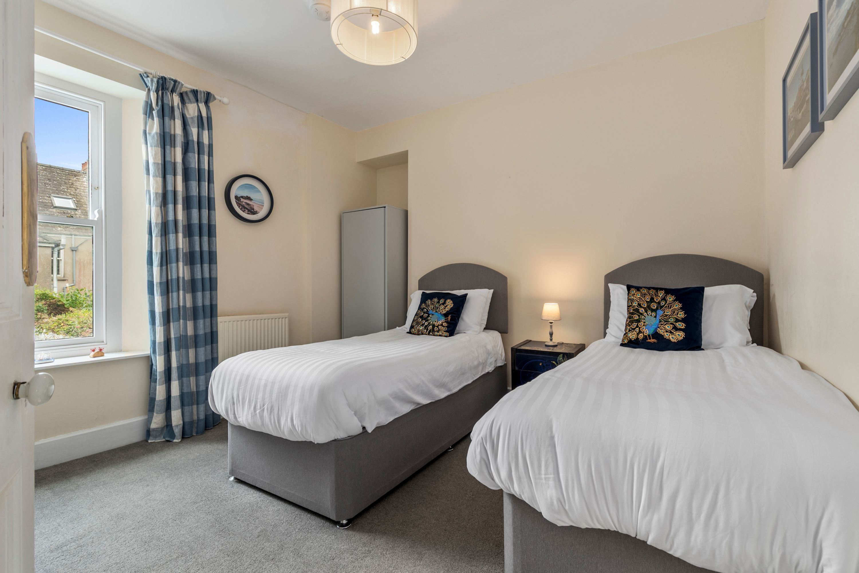 A bedroom with two single beds, a window with curtains, a radiator, a framed picture, and a bedside table with a lamp.