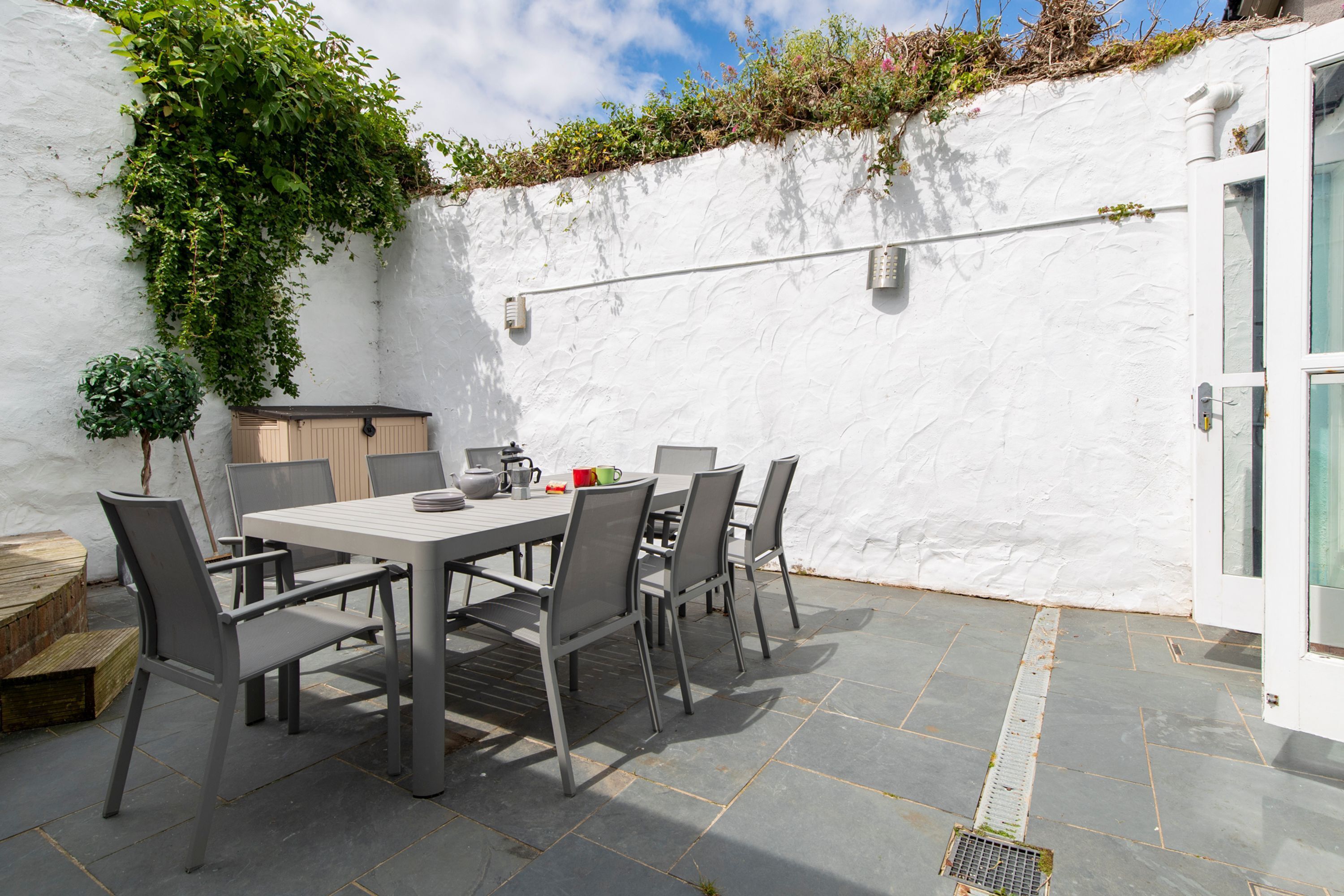 A patio with a table and chairs, a small wooden structure, and a white wall with greenery.