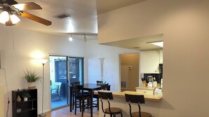 A modern living room with a sofa, a dining table with chairs, a kitchen area with a counter and stools, and a ceiling fan.