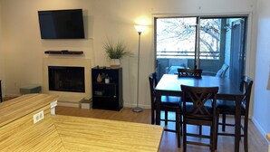 A modern living room with a fireplace, a TV mounted on the wall, a dining table with chairs, and a large window providing a view of the outdoors.