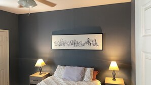 A bedroom with a bed, two bedside tables with lamps, a ceiling fan, and a cityscape artwork on the wall.
