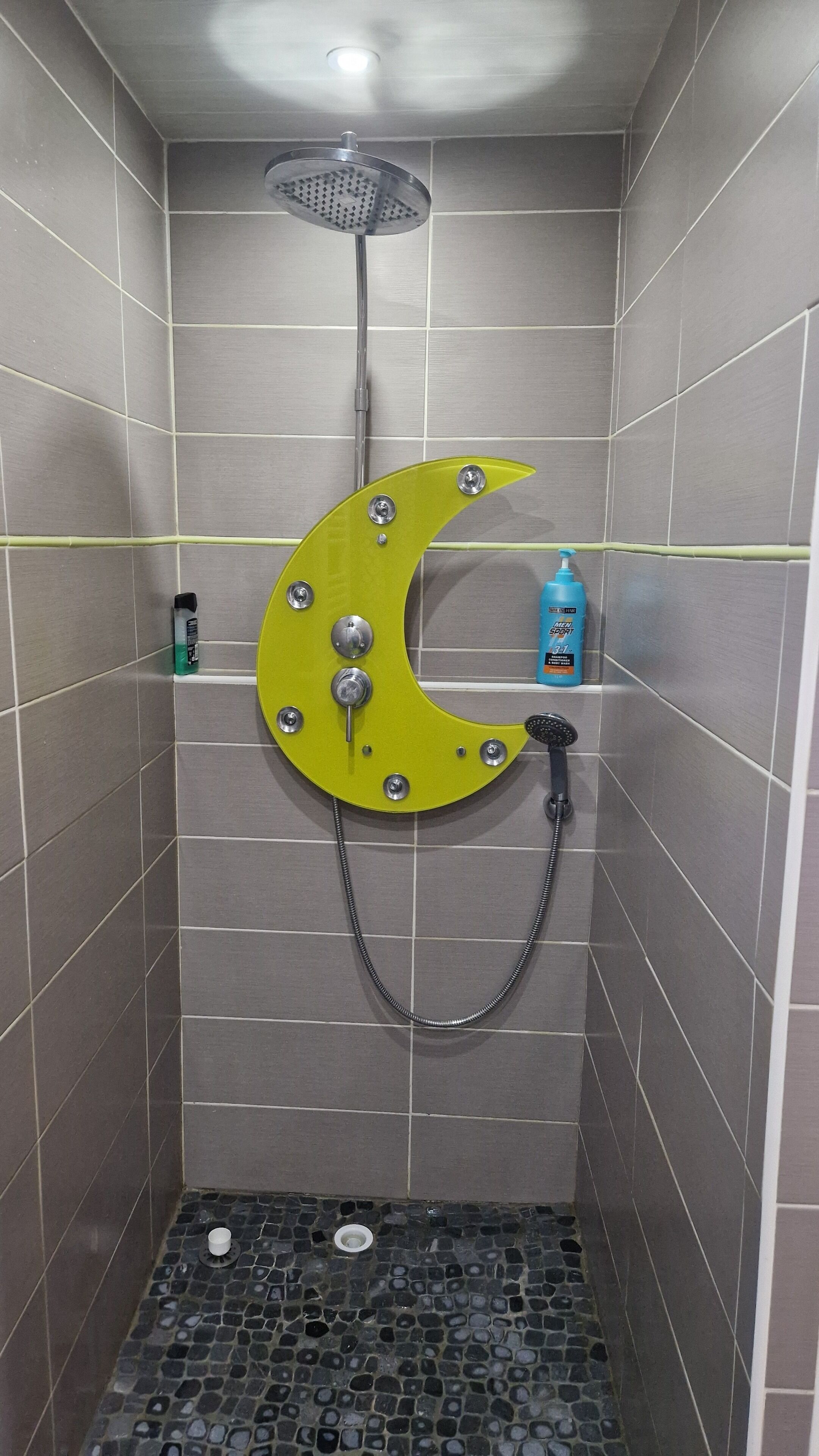 Una ducha con cabezal en forma de luna creciente, una botella de champú deportivo para hombre y un desagüe en el suelo.