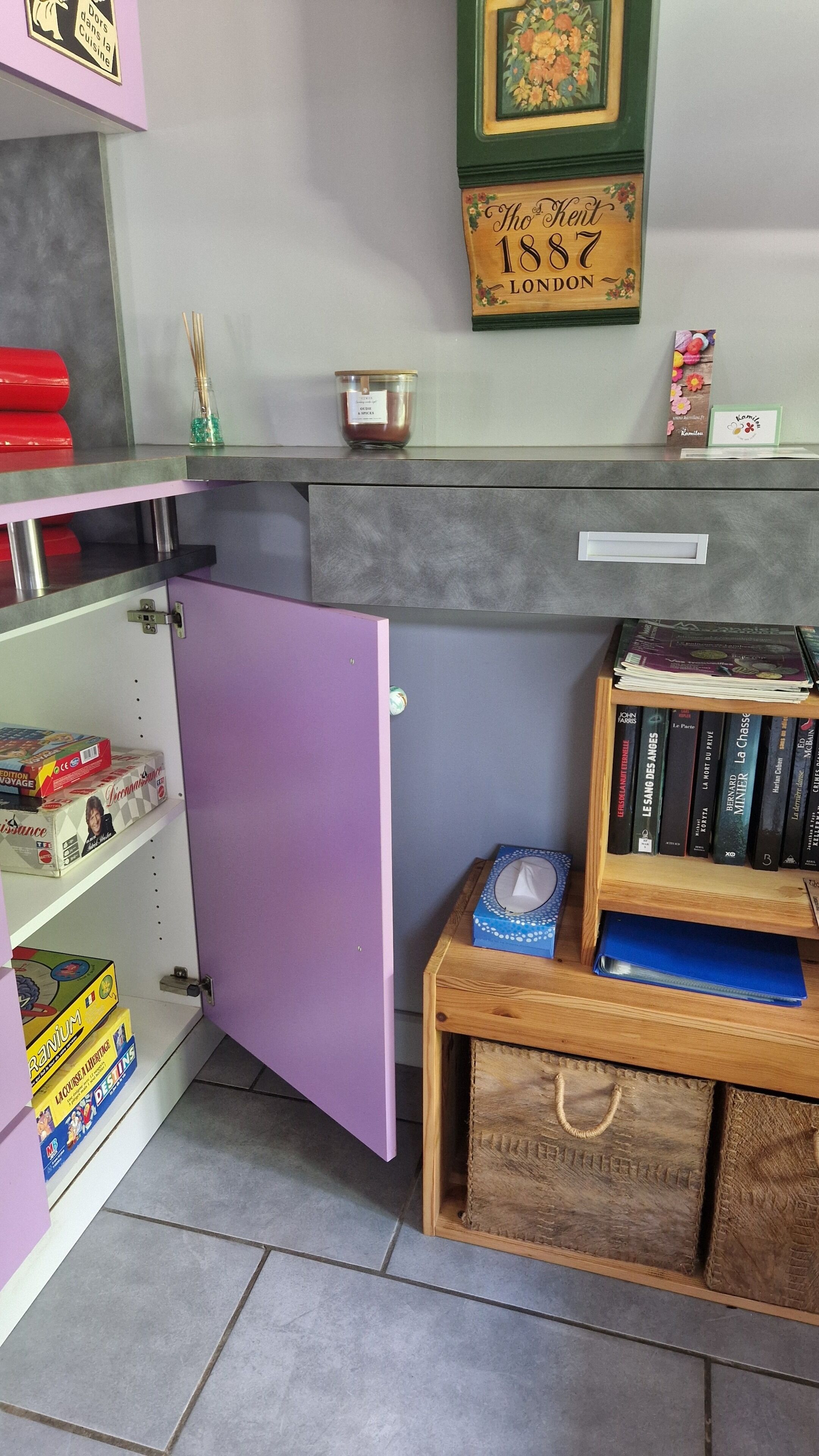 Quarto com um armário roxo, uma pequena mesa de madeira com livros, um quadro com flores e uma estante com diversos objetos.
