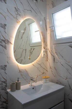 Un bagno con parete in marmo, uno specchio rotondo con illuminazione sottostante, un lavandino bianco e una finestra sopra lo specchio.