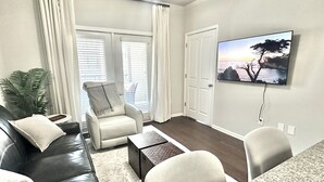 A living room with a black leather sofa, a white armchair, a coffee table, and a TV mounted on the wall.