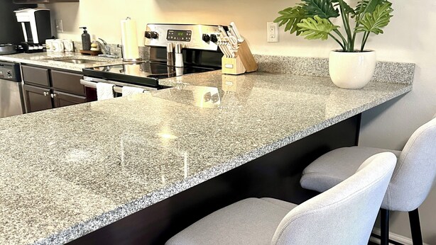 A kitchen with dark cabinets, a granite countertop, a microwave, a stove, and a sink. There are three bar stools and a potted plant on the countertop.