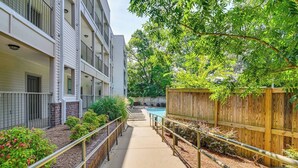 A pathway leading to a pool with a wooden fence and greenery on one side, and a building with balconies on the other.