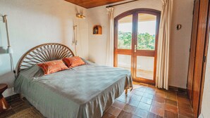 Une chambre avec un plafond en bois, un lit recouvert d’une couverture grise, deux oreillers orange, une table de chevet et une fenêtre avec des rideaux.