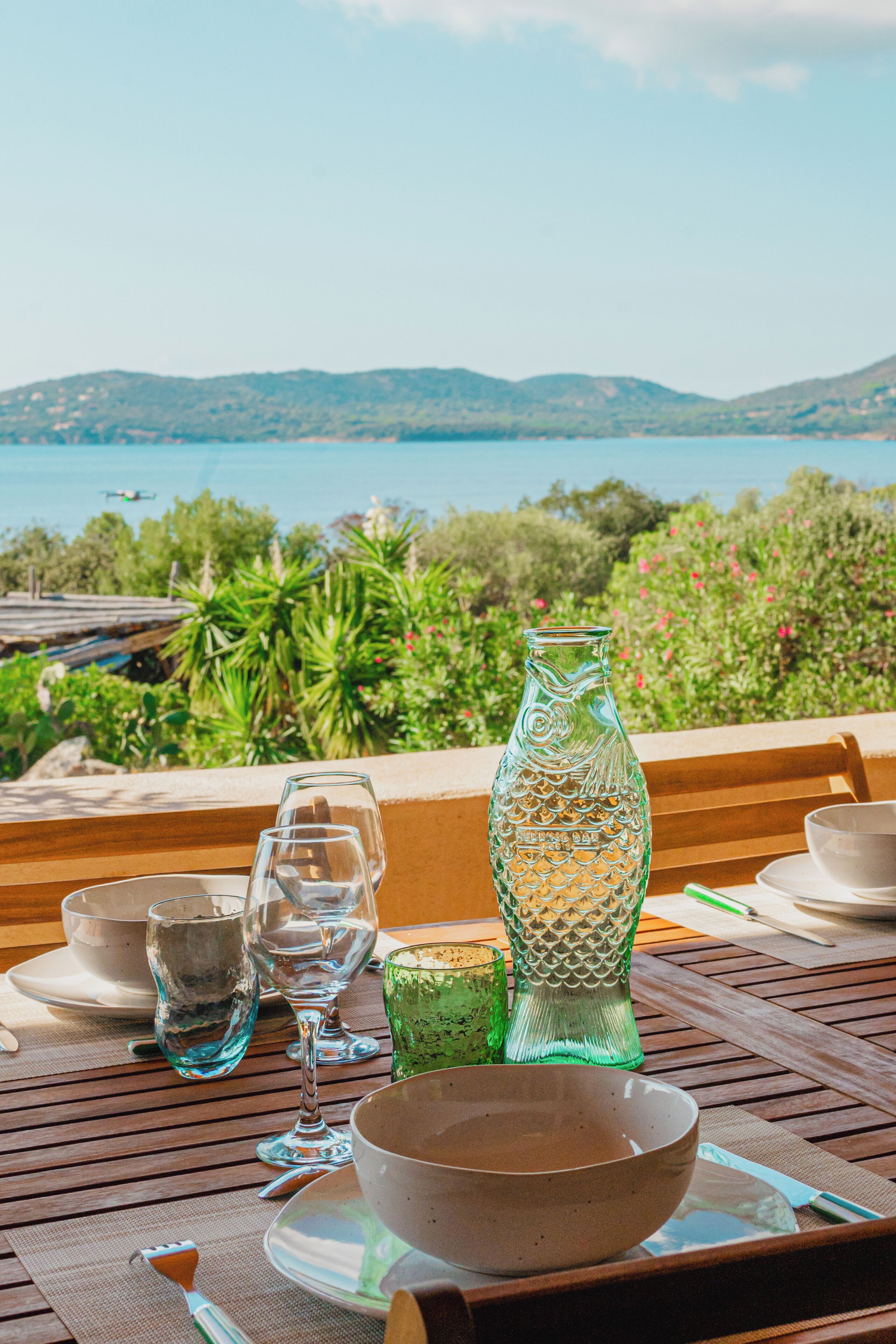 A table set with dinnerware, glasses, and a decorative vase, overlooking a scenic landscape with hills and a body of water.
