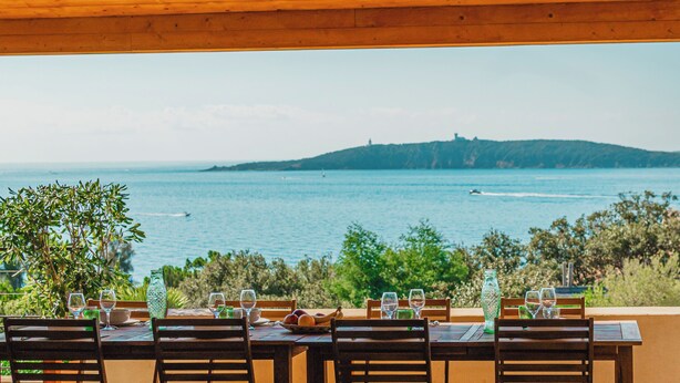 Un espace de restauration en plein air avec une table dressée pour quatre personnes, offrant une vue sur une étendue d’eau et une colline lointaine.