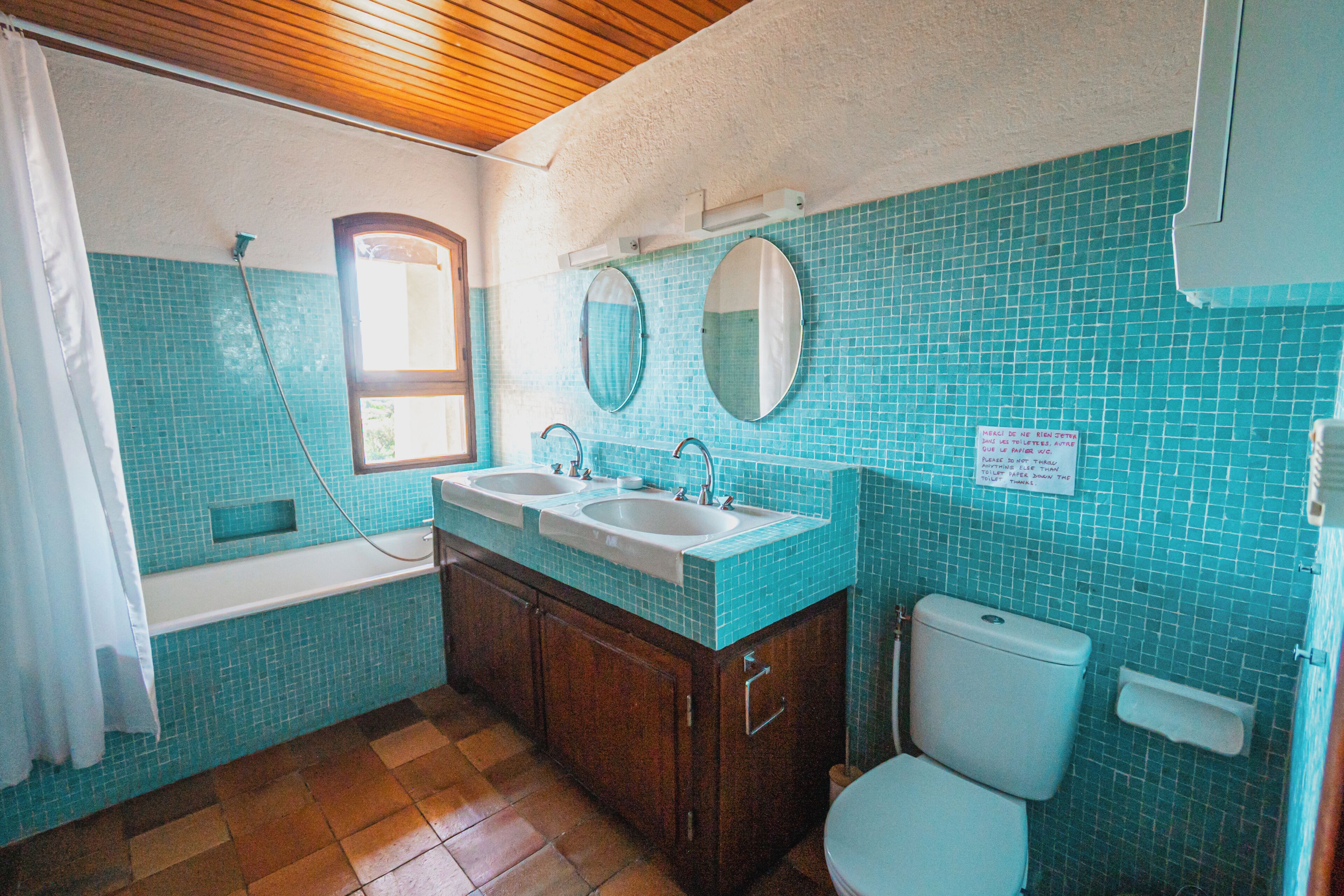 A bathroom with turquoise tiles, a wooden ceiling, a bathtub, two sinks, and a toilet.