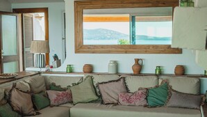 Un salon chaleureux avec un canapé agrémenté de différents coussins, une fenêtre à cadre en bois donnant sur un paysage pittoresque et une étagère ornée d’objets décoratifs.