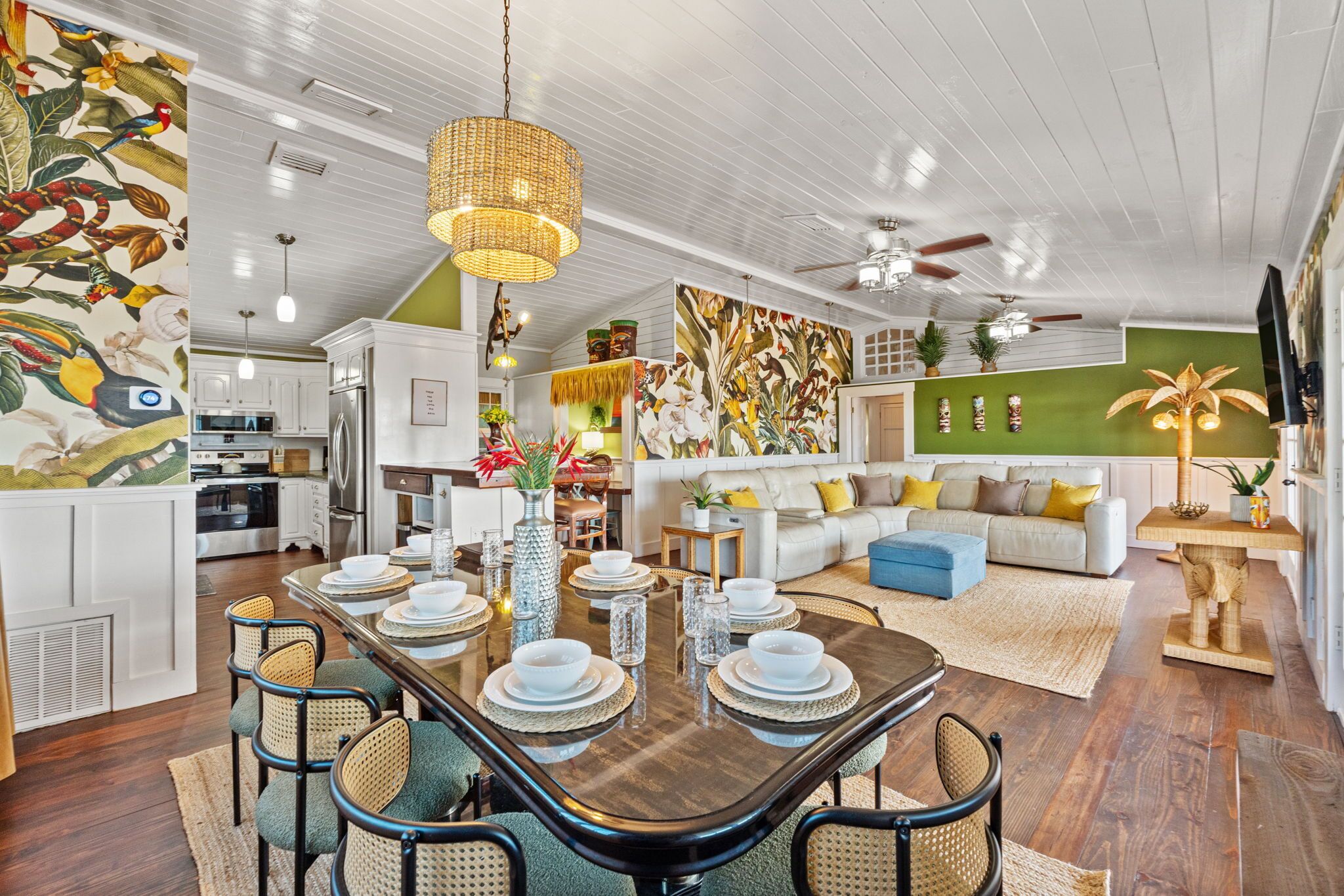 A spacious living area with a large dining table set for six, surrounded by wicker chairs. A kitchen with white cabinets is visible in the background, and a large painting adorns the wall.