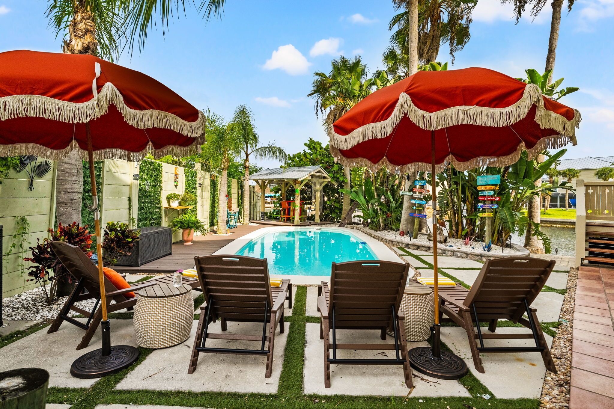 A pool area with lounge chairs and red umbrellas, surrounded by palm trees and a small garden.