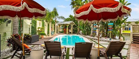 A pool area with lounge chairs and red umbrellas, surrounded by palm trees and a small garden.