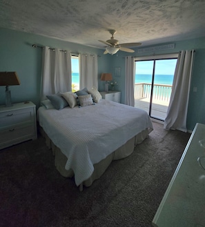 A bedroom with a bed, nightstands, a ceiling fan, and a view of the beach.