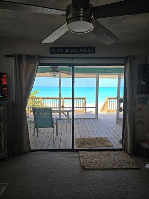 A room with a view of the beach, a ceiling fan, a sign reading 'Nude Beach Ahead', and a wooden deck outside.